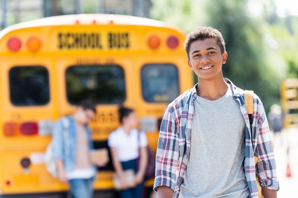 happy teen african american schoolboy looking at camera in front of school bus